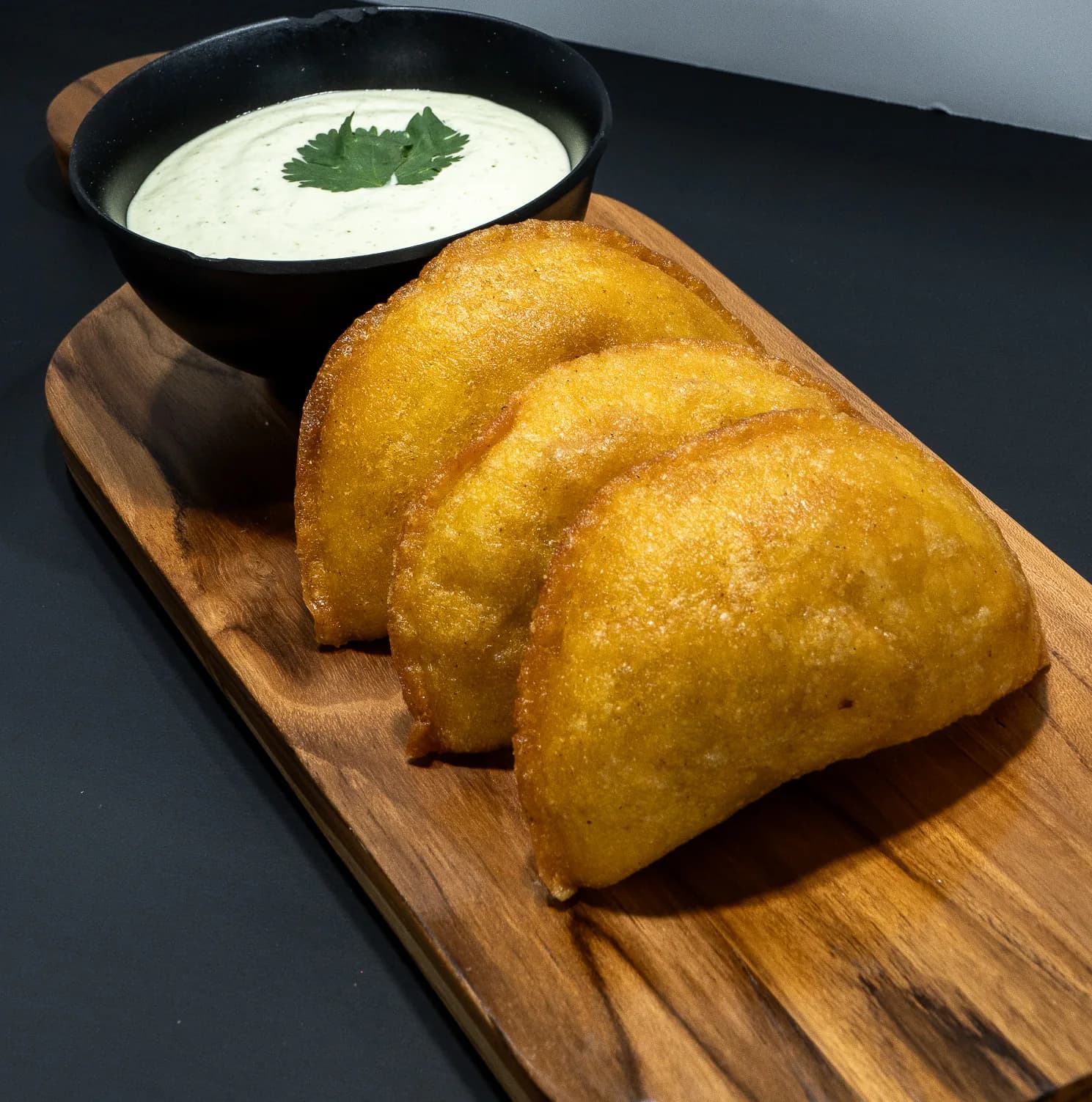 Three golden corn empanadas on a wooden board, with a bowl of creamy sauce